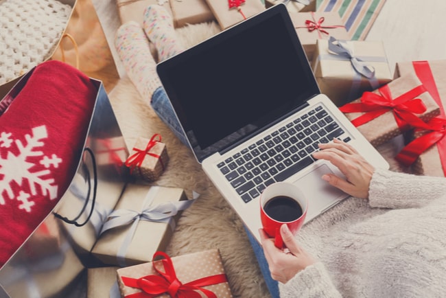Kvinna som sitter med laptop i knät, kaffekopp i handen och julklappar runt omkring sig och handlar julklappar online.