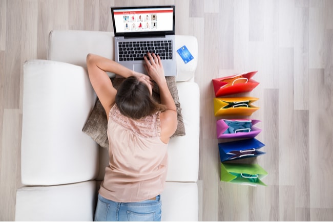Ung kvinna ligger på mage i soffan med laptop framför sig och gör inköp på nätet.